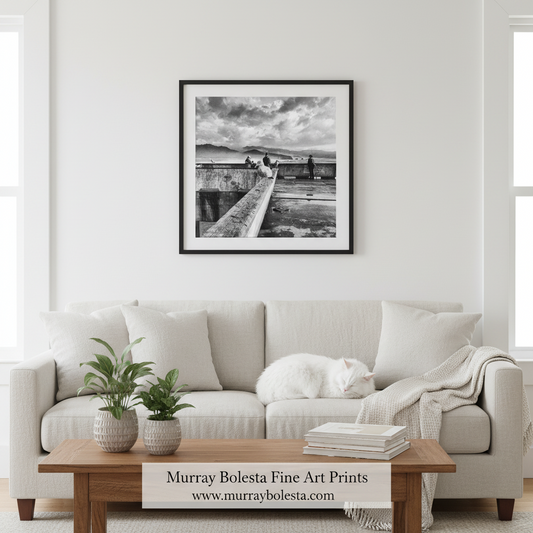 Pacifica Pier. Murray Boletsa. A black and white framed art print of a coastal stone walkway, shown hanging on a white wall above a light beige sofa in a bright living room. A white cat rests on the sofa, and a wooden coffee table with books and potted plants sits in front.