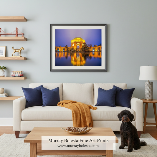 A framed wall art print of a golden illuminated arch reflected in water, displayed above a white sofa in a styled living room with a dog, shelves, and a lamp. The print is branded with a watermark at the bottom.