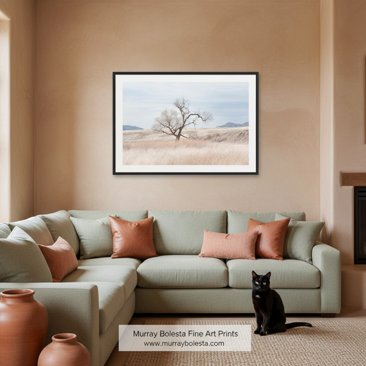 A framed art print depicting a single bare tree in a dry grassy field under a pale sky, displayed on a beige wall above a light sage green sectional sofa with terracotta pillows. A black cat sits on the rug in the foreground, and a white label at the bottom reads 'Murray Bolesta Fine Art Prints 