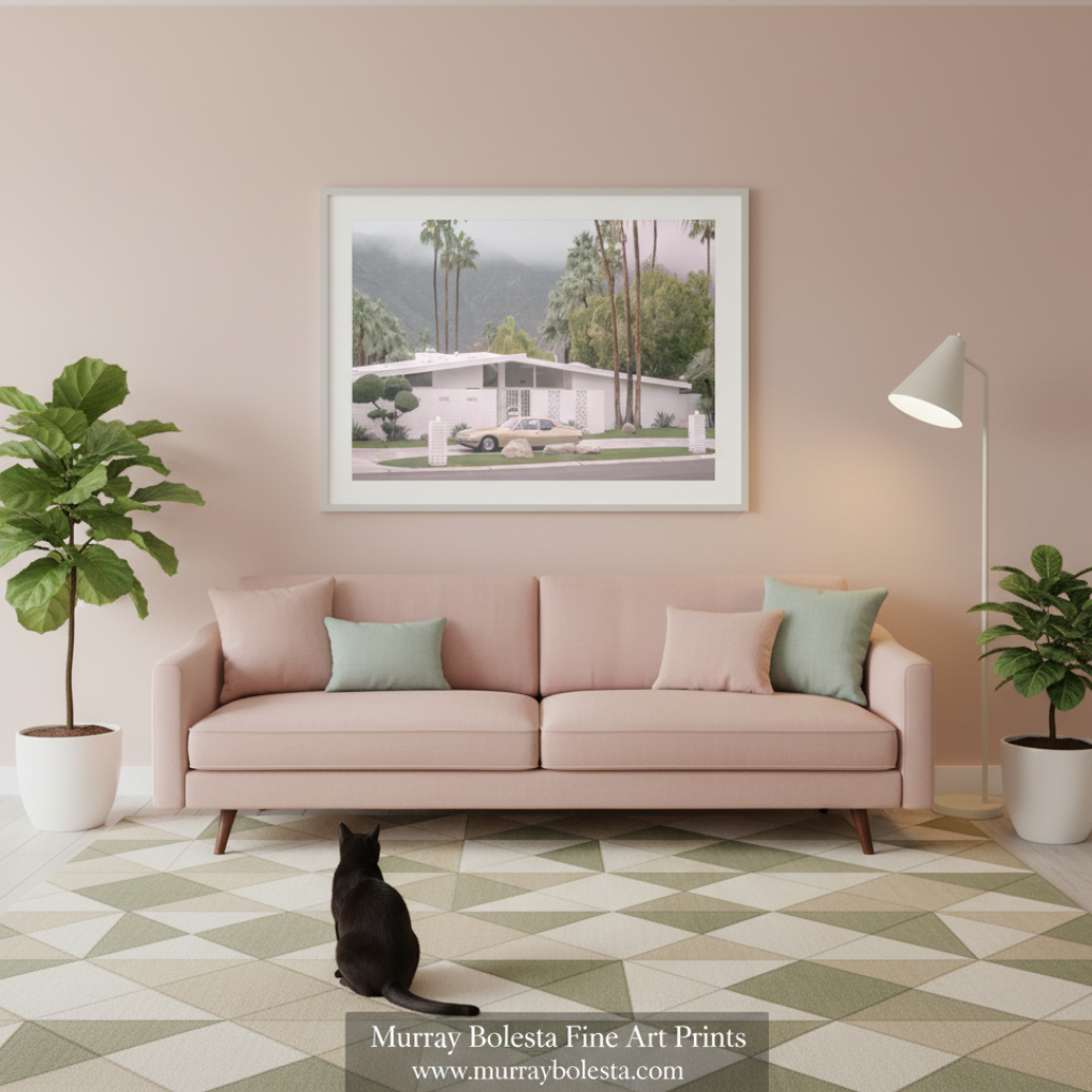 Palm Springs by Murray Bolesta. A framed art print depicting a mid-century modern house with palm trees, displayed on a beige wall above a pink sofa in a styled living room. 