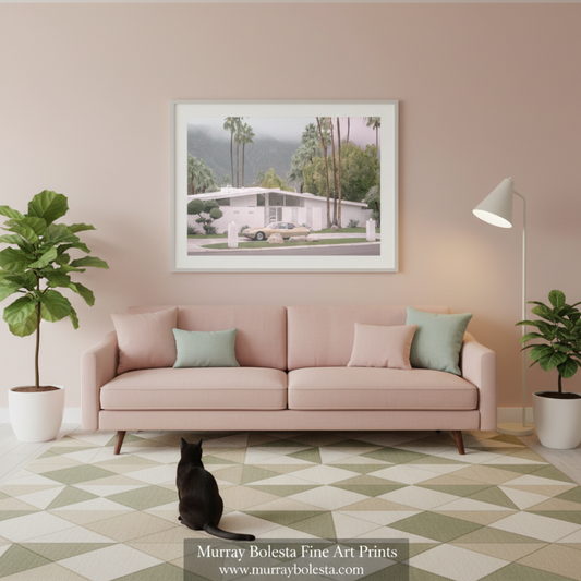 Palm Springs by Murray Bolesta. A framed art print depicting a mid-century modern house with palm trees, displayed on a beige wall above a pink sofa in a styled living room. 