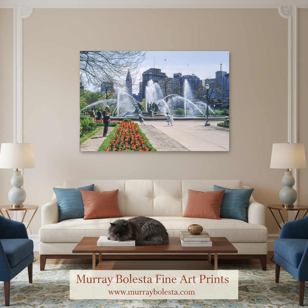 A framed wall art print depicting a city park fountain scene with people and flowers, displayed above a cream sofa in a styled living room. The print is the product being sold, and the room serves as a lifestyle mockup.