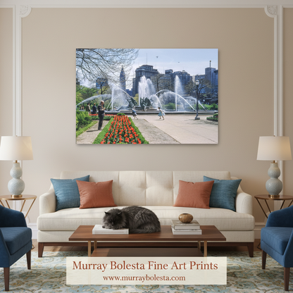 A framed wall art print depicting a city park fountain scene with people and flowers, displayed above a cream sofa in a styled living room. The print is the product being sold, and the room serves as a lifestyle mockup.