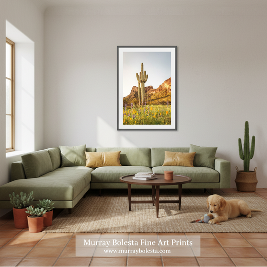 A framed art print of a desert landscape with a saguaro cactus and wildflowers, displayed on a light gray wall above a green sectional sofa in a styled living room. A golden retriever is lying on the rug on the floor.
