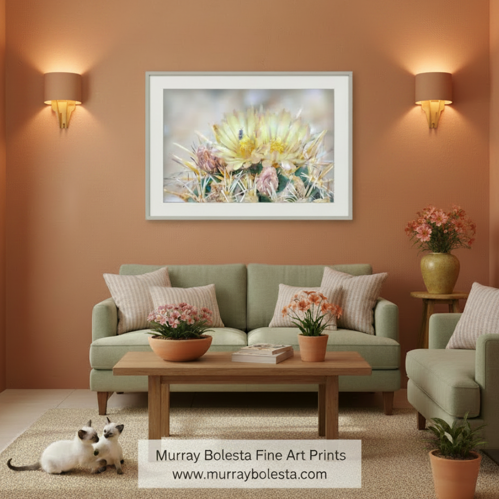A framed floral art print featuring a yellow and pink cactus flower hangs on a terracotta wall above a green sofa in a styled living room. The print is the product being sold, but the image is a lifestyle mockup showing the artwork in a real interior setting with plants, furniture, and a cat.