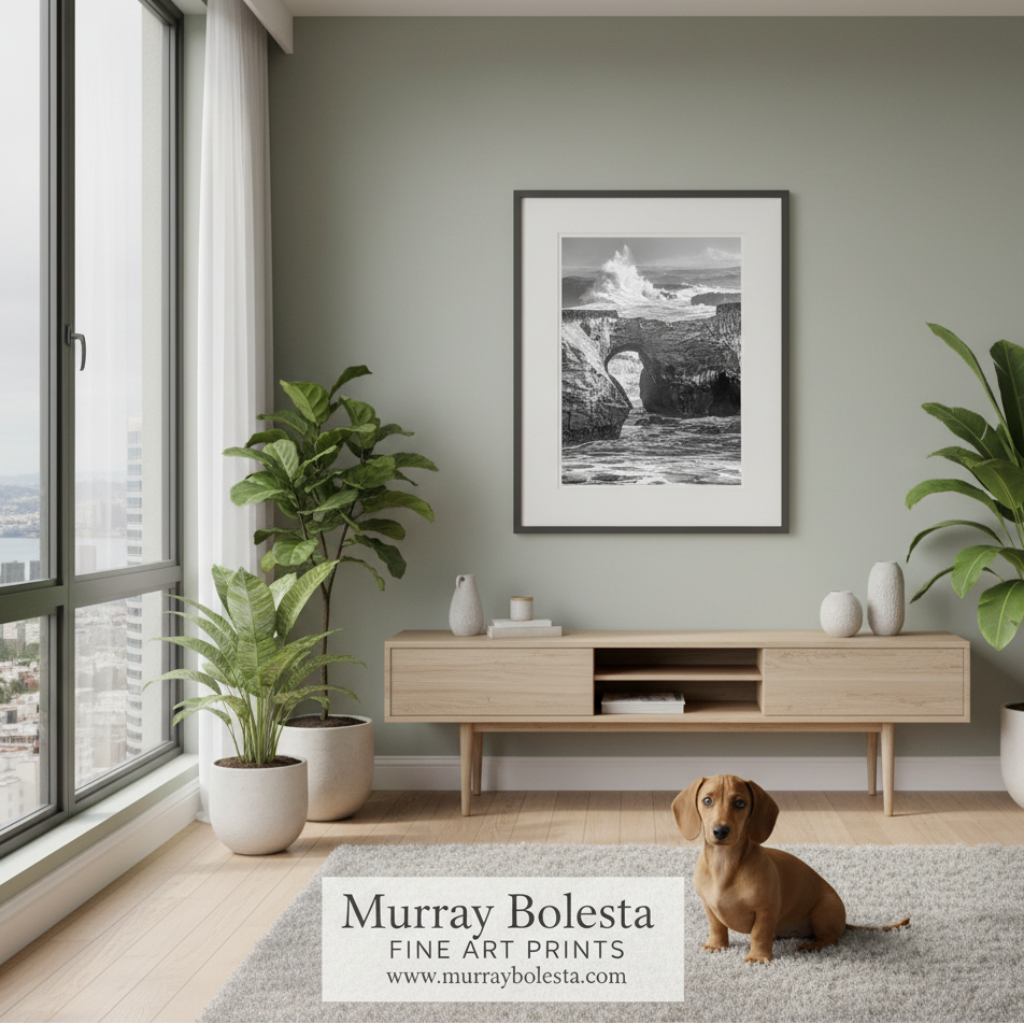 A framed black and white fine art print of a coastal rock formation hangs on a sage green wall in a modern living room. A small brown dachshund sits on a light gray rug in front of a light wood console table, with potted plants and a large window in the background.