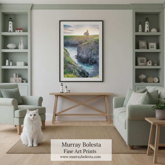 A framed vertical art print of a lighthouse on a rocky cliff by Murray Bolesta hangs on a light gray wall in a styled living room. A white cat sits on the floor in front of the console table, flanked by two pale green armchairs and built-in shelving.
