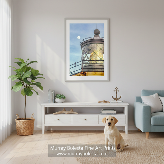 A framed art print of a lighthouse under a blue sky with a moon, displayed on a light gray wall in a styled living room above a white console table with a yellow Labrador sitting in front. A potted plant is on the left, and a blue armchair is on the right.