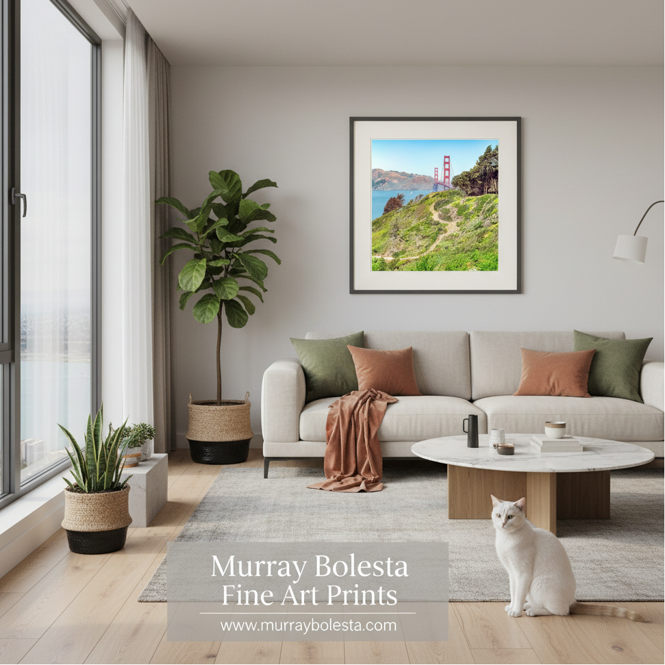 Presidio Bluff by Murray Bolesta.A framed art print of the Golden Gate Bridge and Marin Headlands hanging on a light gray wall in a modern living room with a beige sofa, plants, and a white cat. Text at the bottom reads 'Murray Bolesta Fine Art Prints'.
