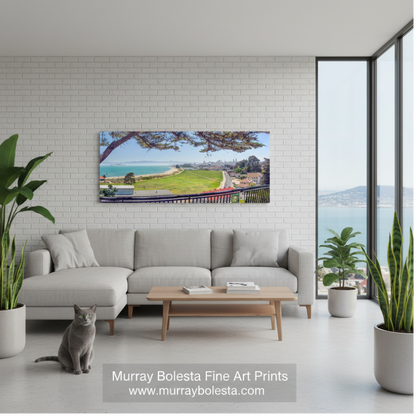 Presidio Chrissy Field! A panoramic landscape art print by Murray Bolesta depicting a coastal green racecourse under a tree, shown hanging on a white brick wall in a modern living room mockup with a gray sectional sofa and a cat.