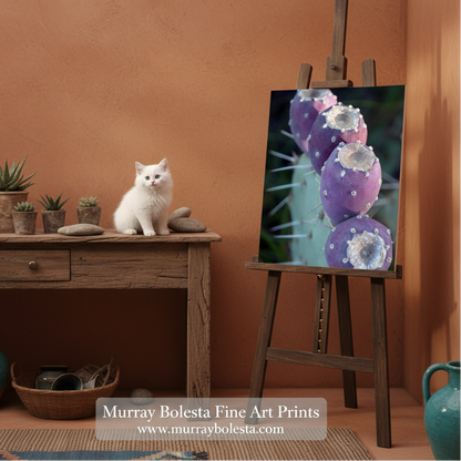 Purple Arizona cactus by Murray Bolesta! A framed-style art print depicting purple cactus fruit with white spines is displayed on a wooden easel in a warm, rustic room. A small white cat sits on a wooden table to the left, and the background features a terracotta-colored wall and decorative plants.