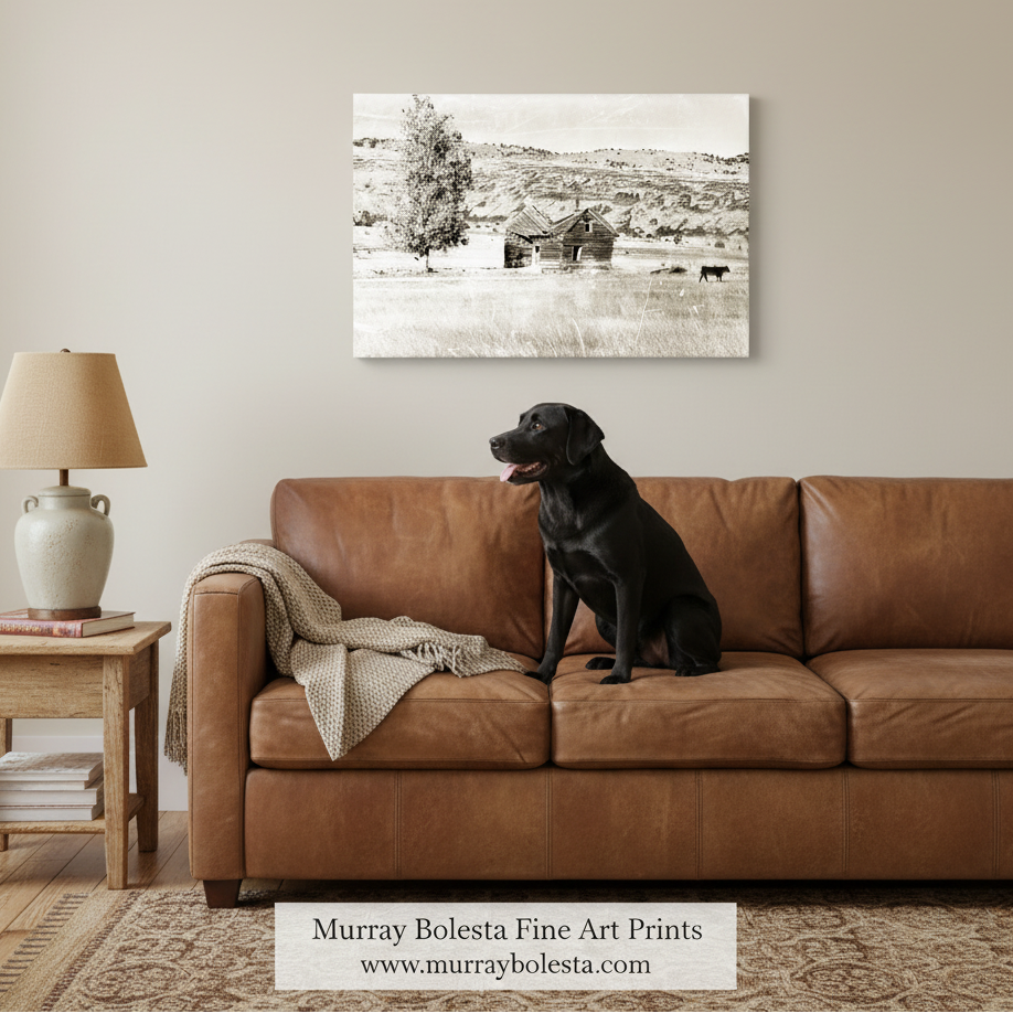 Ranch log cabin in northeast California. A black and white landscape art print by Murray Bolesta hangs on a beige wall above a brown leather sofa with a black dog sitting on the couch. A side table with a lamp is visible on the left.