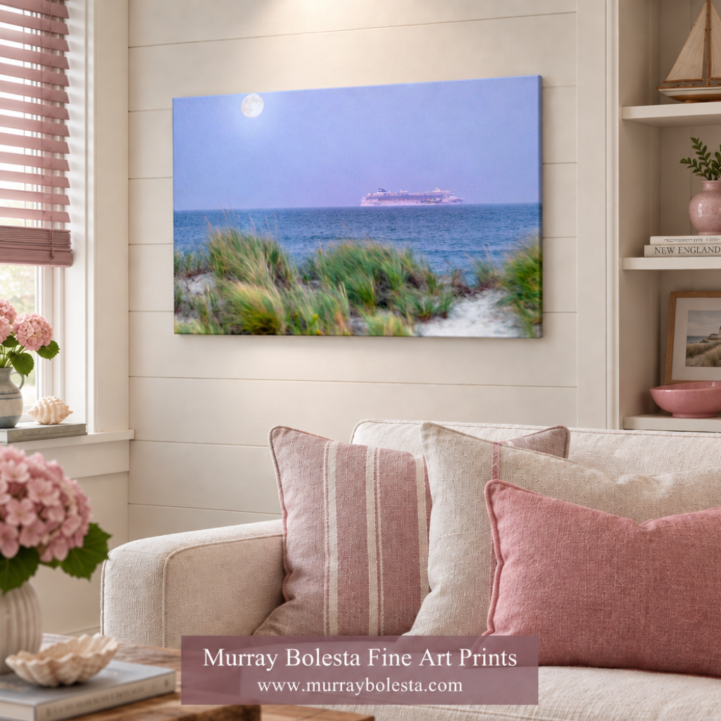 A framed wall art print showing a coastal seascape with dune grasses, blue water, a ship, and a bright moon, displayed above a sofa in a styled living room. A watermark at the bottom reads 'Murray Bolesta Fine Art Prints' with the website.