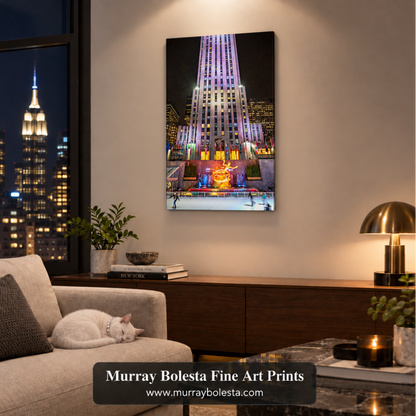 A framed vertical wall art print depicting the illuminated Rockefeller Center at night hangs on a beige wall in a modern living room. A white cat rests on a light beige sofa to the left, with a wooden console, lit table lamp, and city skyline visible through the window. Art by Murray Bolesta