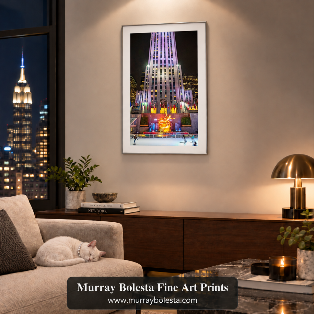 A framed vertical art print of the illuminated Rockefeller Center at night hangs on a beige wall in a modern living room. A white cat rests on a light beige sofa to the left, and a dark wood console table with a brass table lamp sits below. Art by Murray Bolesta