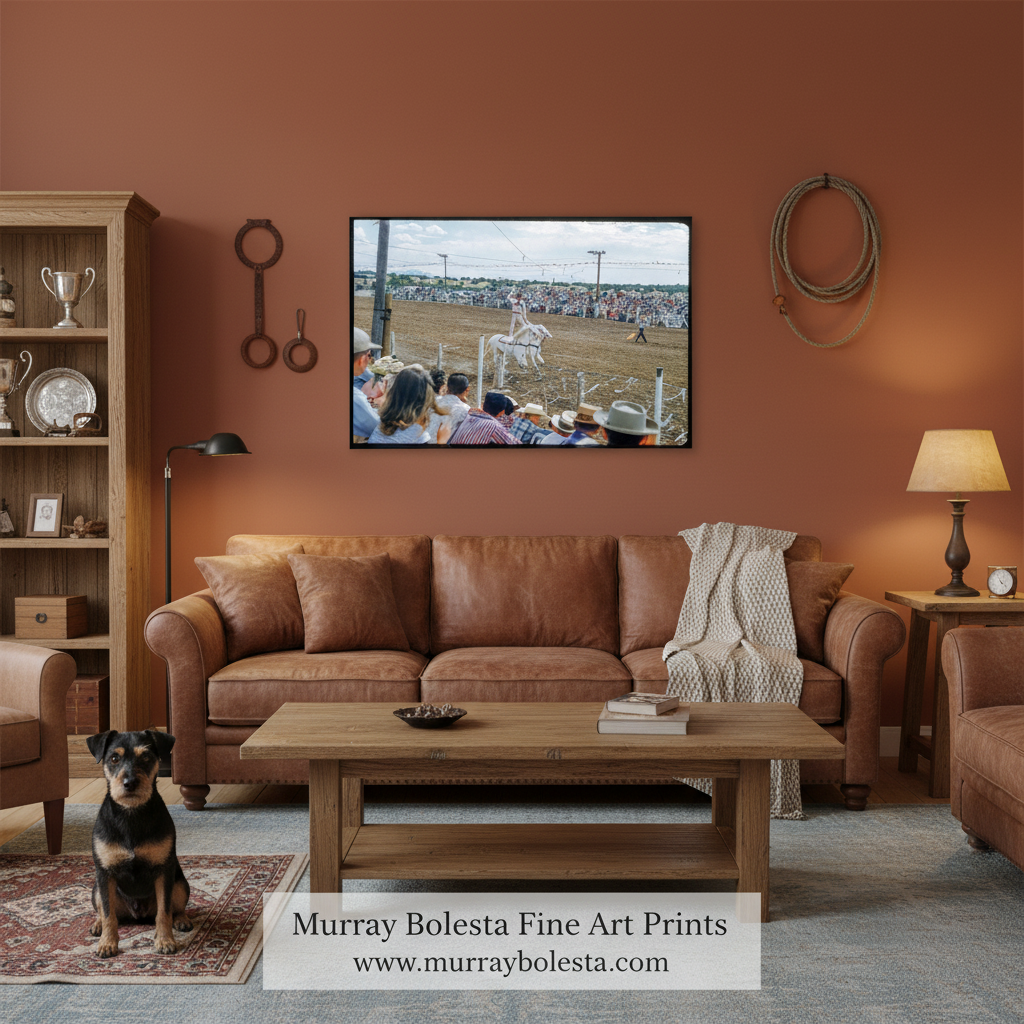 A framed wall art print showing a rustic outdoor scene with people at a rodeo, displayed in a styled living room above a brown leather sofa. The room has warm terracotta walls, a wooden coffee table, a leather chair, and a small dog sitting on the floor.