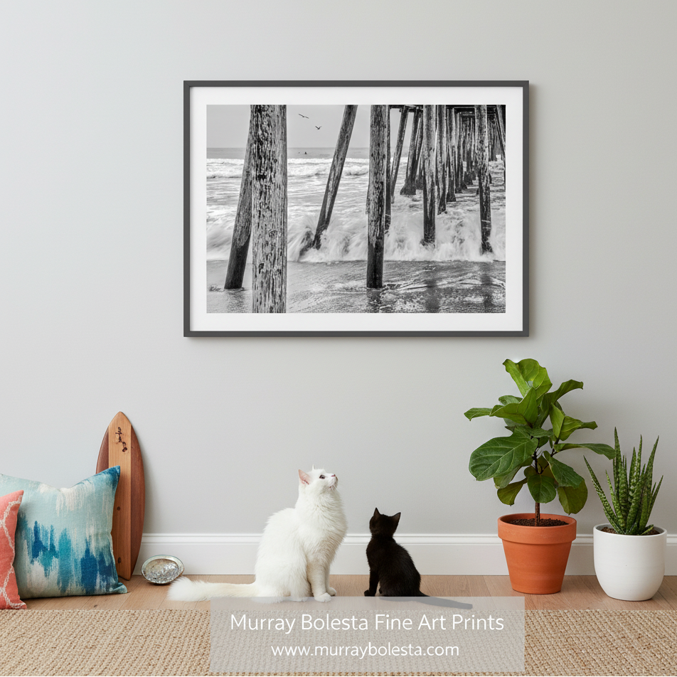 A black and white photograph of a wooden pier extending into the ocean, with waves crashing against the posts and the water churning around them. Imperial Beach Pier Print – San Diego Black & White Art by Murray Bolesta!