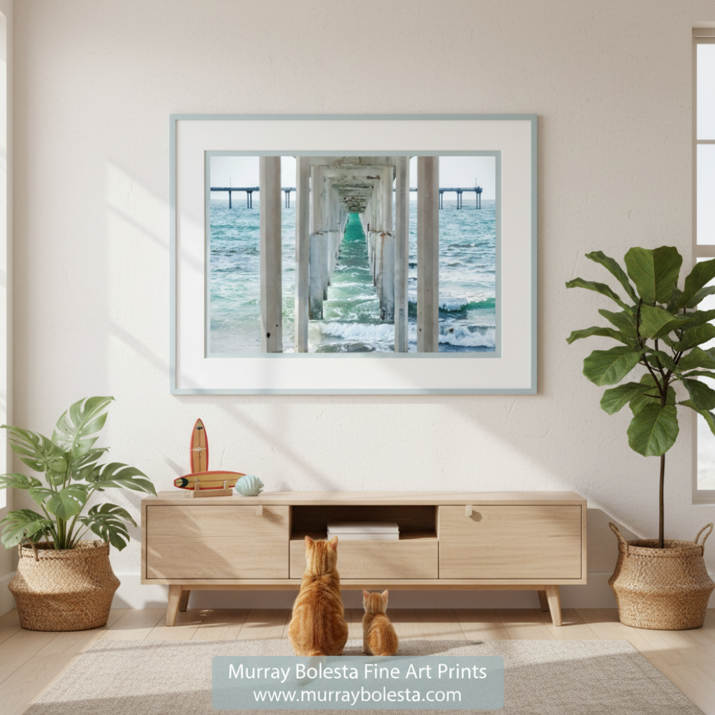 Art by Murray Bolesta! A long, white pier extends into the ocean, with a series of white columns supporting the structure. The water below the pier is a light blue-green color, and the sky above is a pale blue. Ocean Beach Pier Print – San Diego Pacific Coast Wall Art