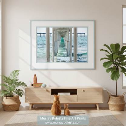 Art by Murray Bolesta! A long, white pier extends into the ocean, with a series of white columns supporting the structure. The water below the pier is a light blue-green color, and the sky above is a pale blue. Ocean Beach Pier Print – San Diego Pacific Coast Wall Art