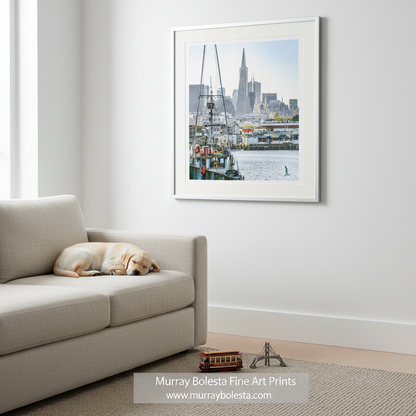 A framed art print of a San Francisco harbor scene with boats and the Transamerica Square tower, displayed on a white wall above a beige sofa with a sleeping dog. By Murray Bolesta!