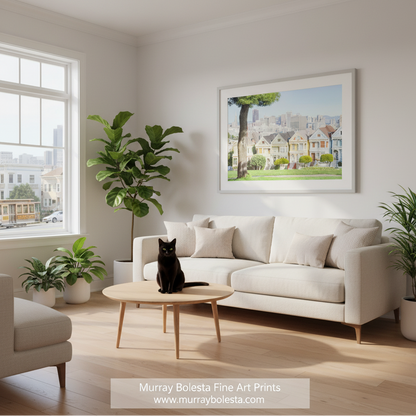 Painted Ladies San Francisco by Murray Bolesta. A framed art print depicting a sunny street scene with colorful Victorian-style houses, a large tree, and a city skyline, displayed on a light gray wall above a cream sofa in a modern living room. A black cat sits on a round wooden coffee table in the foreground.