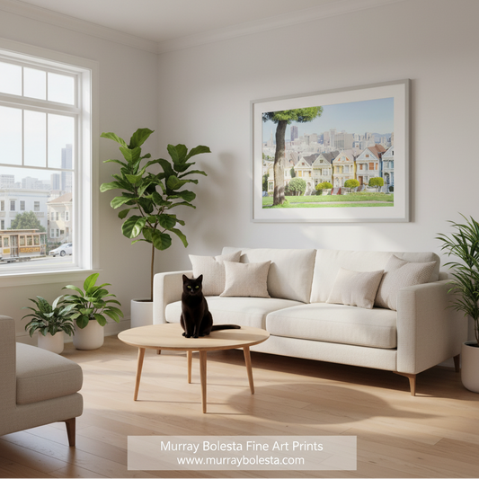 Painted Ladies San Francisco by Murray Bolesta. A framed art print depicting a sunny street scene with colorful Victorian-style houses, a large tree, and a city skyline, displayed on a light gray wall above a cream sofa in a modern living room. A black cat sits on a round wooden coffee table in the foreground.