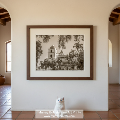 Santa Barbara Mission California by Murray Bolesta! A framed sepia art print of a historic mission building surrounded by palm trees, displayed on a cream wall in a home interior with a white cat sitting below the frame.