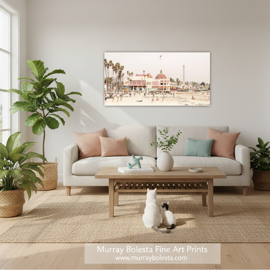 Santa Cruz Beach Boardwalk by Murray Bolesta! A framed art print depicting a beach boardwalk scene with palm trees and a building, displayed on a light gray wall above a beige sofa in a styled living room. The room includes a wooden coffee table, potted plants, and a white cat sitting on the rug.