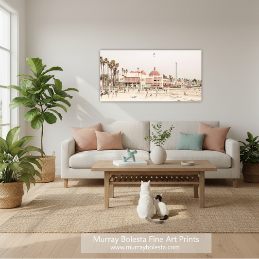 Santa Cruz Beach Boardwalk by Murray Bolesta! A framed art print depicting a beach boardwalk scene with palm trees and a building, displayed on a light gray wall above a beige sofa in a styled living room. The room includes a wooden coffee table, potted plants, and a white cat sitting on the rug.