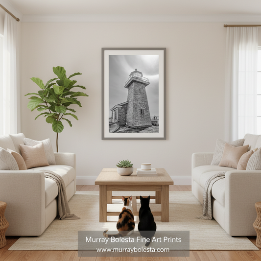 Santa Cruz Surfer's Lighthouse art print! A framed black and white art print of a lighthouse hanging on a beige wall in a modern, bright living room with two cream sofas, a wooden coffee table, and two cats sitting on the floor. A watermark at the bottom reads 'Murray Bolesta Fine Art Prints'.