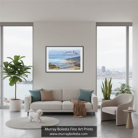 A framed landscape art print by Murray Bolesta hangs on a light gray wall above a modern beige sofa in a bright, contemporary living room. The print depicts a coastal city and sea scene.