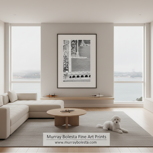 A framed black and white fine art print of a bridge hangs on a light gray wall in a modern, sunlit living room with a beige sectional sofa, a small round coffee table, and a white dog lying on a gray rug. A watermark at the bottom reads 'Murray Bolesta Fine Art Prints'.