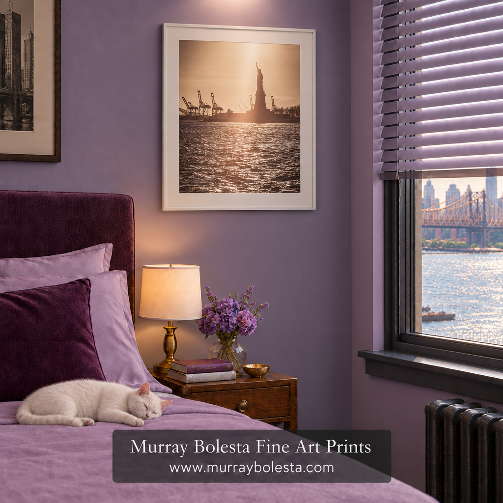 A framed art print of the Statue of Liberty at sunset hangs on a lavender-purple bedroom wall above a bed with purple bedding and a sleeping white cat. A window with blinds shows a city waterfront view, and a text banner at the bottom reads 'Murray Bolesta Fine Art Prints www.murraybolesta.com