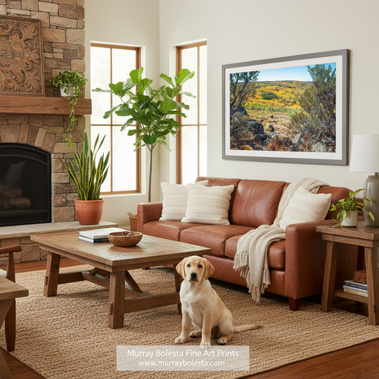 Steens Mountain Oregon. A framed landscape art print by Murray Bolesta hangs on a beige wall in a cozy living room. The print depicts a sunny yellow and green hillside with rocky terrain under a blue sky.