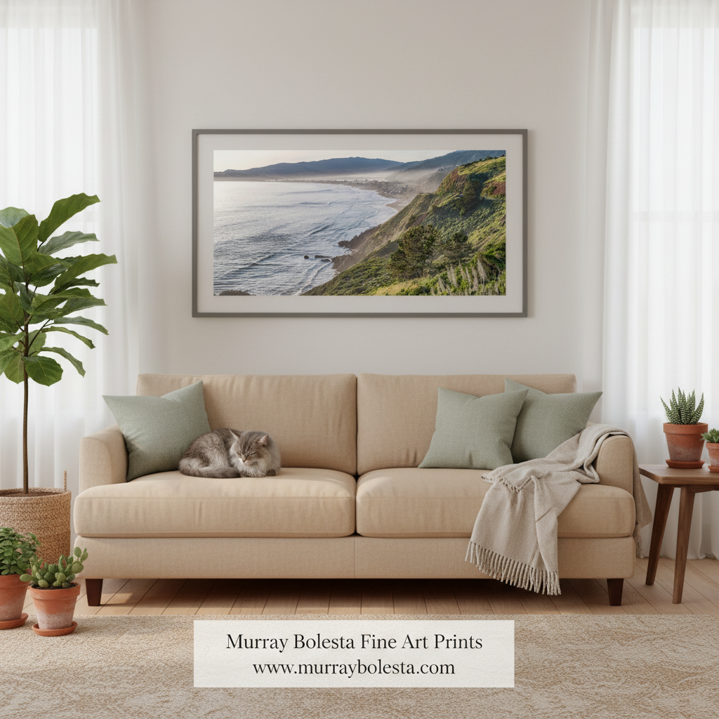 Stinson Beach California. A framed landscape art print of a coastal cliffside ocean scene hangs on a beige wall above a cream sofa with green pillows and a throw blanket in a styled living room. A cat rests on the sofa, and a brand watermark at the bottom reads 'Murray Bolesta Fine Art Prints'.