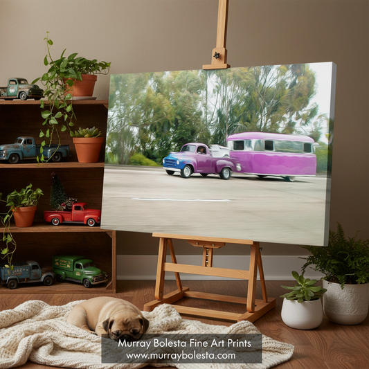 studebaker truck by Murray Bolesta. A canvas art print displayed on a wooden easel in a styled room. The print depicts a vintage purple camper trailer and blue pickup truck on a road with trees in the background.