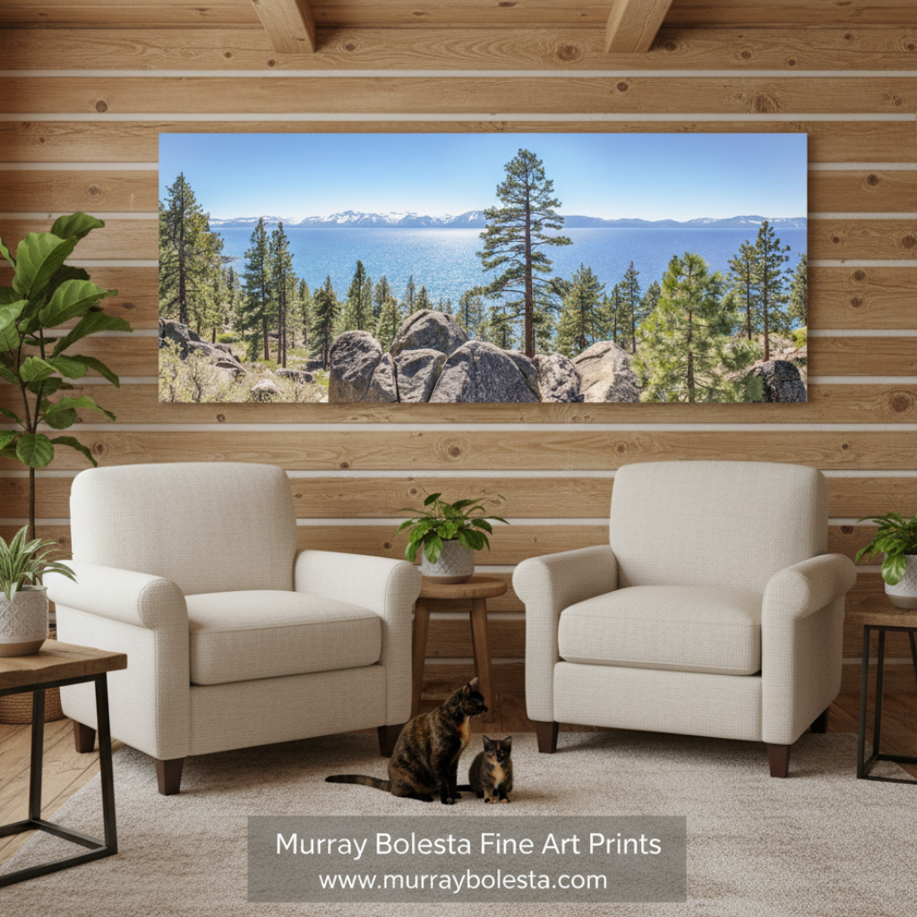 Tahoe art panorama by Murray Bolesta! A large rectangular wall art print showing a scenic lake and pine forest landscape, displayed above two cream armchairs in a wood-paneled living room. A cat sits on the rug in front of the chairs.