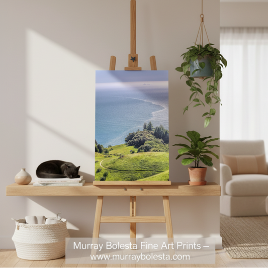 Mount Tamalpais California by Murray Bolesta! A vertical fine art print of a coastal landscape with green hills and blue ocean, displayed on a wooden easel in a styled room setting with a potted plant, a sleeping cat, and a sofa in the background.