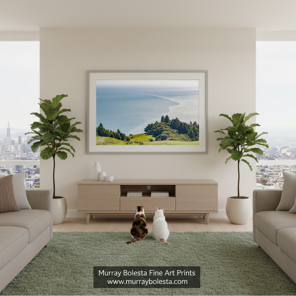 Mount Tamalpais and Farallon Islands by Murray Bolesta! A framed landscape art print of a coastal bay and green hills hangs on a beige wall in a modern living room. Two cats are sitting on the floor in front of the console, flanked by two potted fiddle-leaf fig plants.