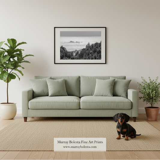 A framed black and white landscape art print hangs on a beige wall above a light sage green sofa in a styled living room. A dachshund sits on the beige rug in front of the sofa, with a potted fiddle-leaf fig on the left and another plant on the right.