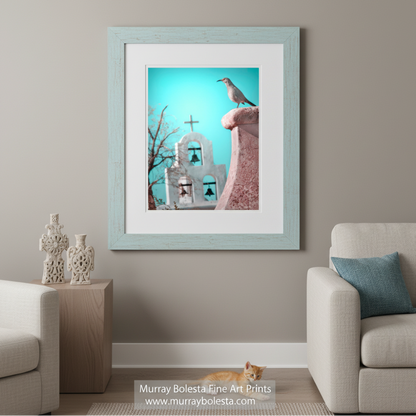 A bird perches on a pink stone structure, with a white church featuring a cross and bell in the background. Surreal Bird Art | San Xavier Mission Chapel Tucson Arizona Print by Murray Bolesta