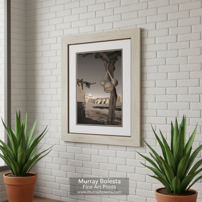 A weathered wooden post with a large, round object hanging from it, set against a backdrop of a desert-like landscape with a domed building in the distance. Tumacacori Mission Historic Church Print - Southwest Gourd Art by Murray Bolesta
