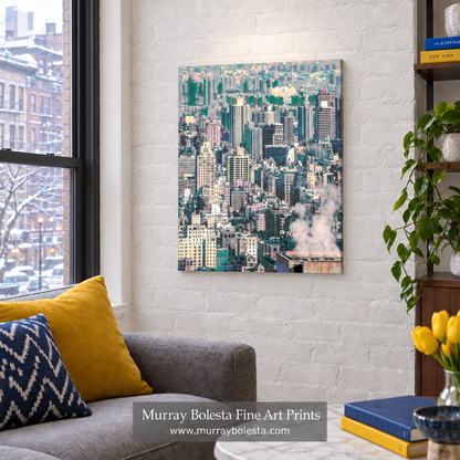 A vertical canvas art print of a dense city skyline with green trees and white steam, displayed on a white brick wall in a modern living room setting next to a gray sofa and yellow pillows. NYC winter art by Murray Bolesta