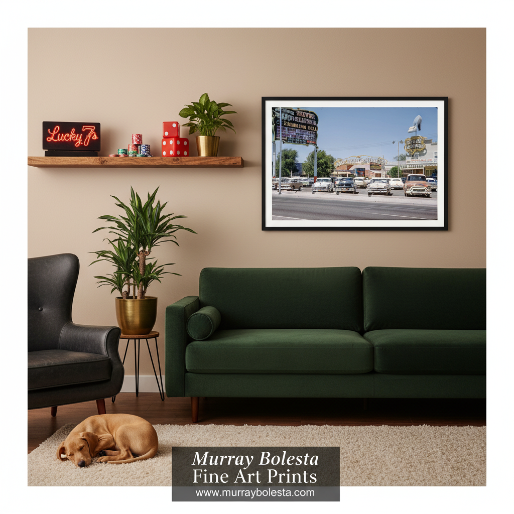 Las Vegas vintage. A framed fine art print of a street scene with vintage cars and a sign, displayed on a beige wall in a styled living room above a dark green sofa. The room includes a wooden shelf with decor, a potted plant, a black armchair, a sleeping dog on a rug, and a watermark at the bottom reading 'Murray Bolesta Fine Art Prints'.