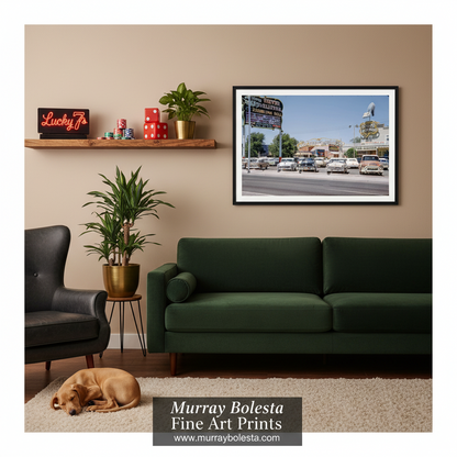 Las Vegas vintage. A framed fine art print of a street scene with vintage cars and a sign, displayed on a beige wall in a styled living room above a dark green sofa. The room includes a wooden shelf with decor, a potted plant, a black armchair, a sleeping dog on a rug, and a watermark at the bottom reading 'Murray Bolesta Fine Art Prints'.