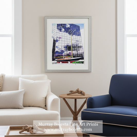 Kennedy Victura sailboat. A framed art print featuring a colorful photograph of a modern glass skyscraper reflecting blue sky and trees, styled in a bright living room above a cream sofa and a navy blue armchair. By Murray Bolesta.