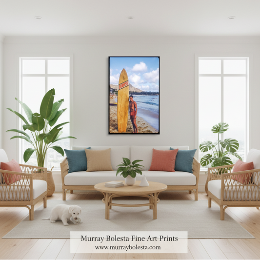 A framed vertical art print of a surfer holding a surfboard on a beach hangs on a white wall above a light beige sofa in a bright, modern living room. The print features a surfer in a red jacket holding a yellow surfboard with ocean and mountain scenery in the background.