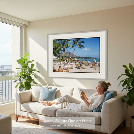 A framed beach scene art print hangs on a beige wall above a light-colored sofa in a bright living room. A woman reclines on the sofa reading a book.