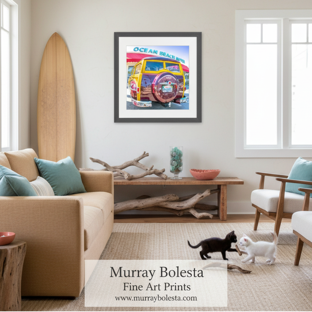 A framed art print titled 'Ocean Beach' featuring a colorful vintage beach car, displayed on a beige wall in a modern, sunlit living room with a sofa, chairs, and two cats on a woven rug.