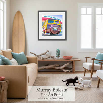 A framed art print titled 'Ocean Beach' featuring a colorful vintage beach car, displayed on a beige wall in a modern, sunlit living room with a sofa, chairs, and two cats on a woven rug.